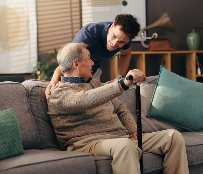 Caregiver assisting elderly man with cane on couch