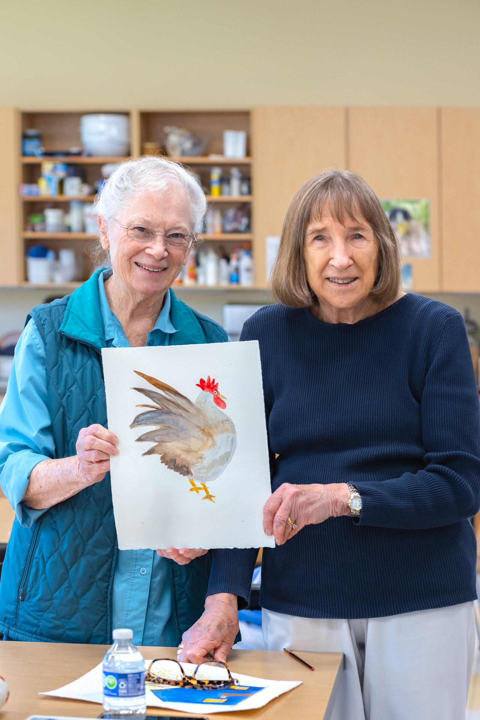 Two elderly women holding rooster painting indoors